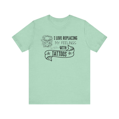 A sarcastic graphic tee stating 'I love replacing my feelings with tattoos' in color heather mint